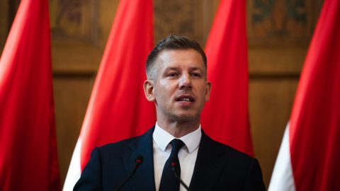 Man in a dark suit and tie speaks at a podium with red banners behind him.