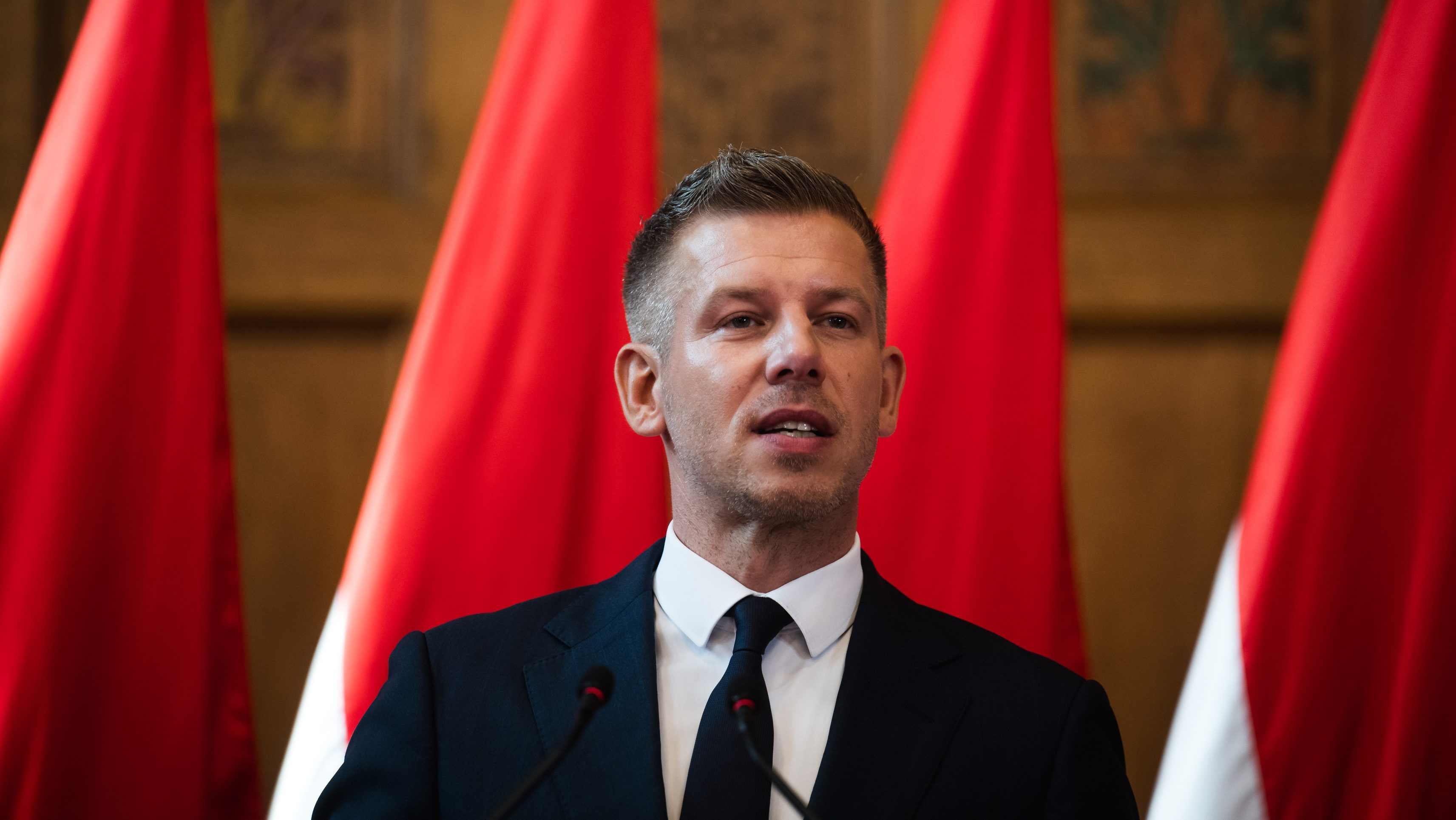 Man in a dark suit and tie speaks at a podium with red banners behind him.
