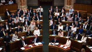 Politicians in formal attire seated in a wood-paneled parliamentary chamber, viewed from above.