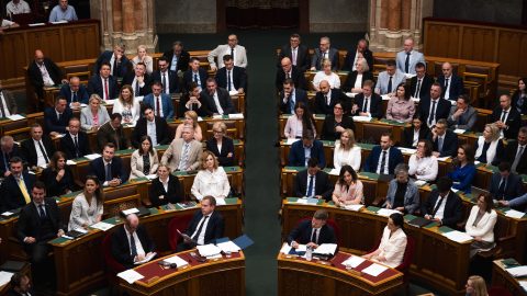 Politicians in formal attire seated in a wood-paneled parliamentary chamber, viewed from above.