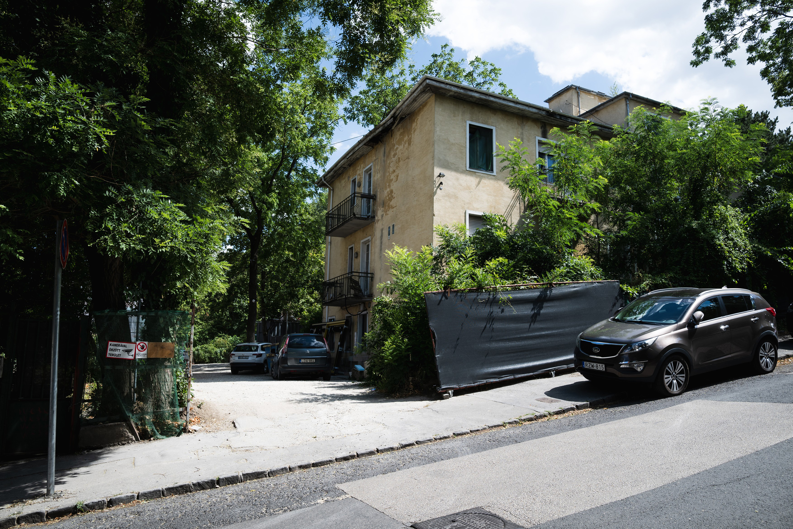 Beige multi-story apartment building partially hidden by dense green trees, with parked cars along the street in front of it.