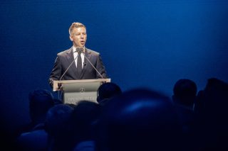 Man in a dark suit and tie speaks into microphones at a lectern on a deep blue stage.