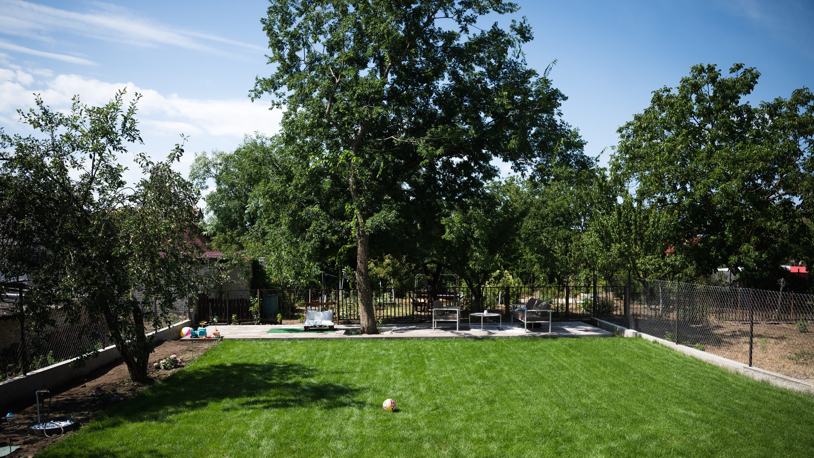 Backyard scene with a green lawn, several trees, a chain-link fence, and patio seating on a concrete area in the background, plus a white robotic lawn mower on the grass.