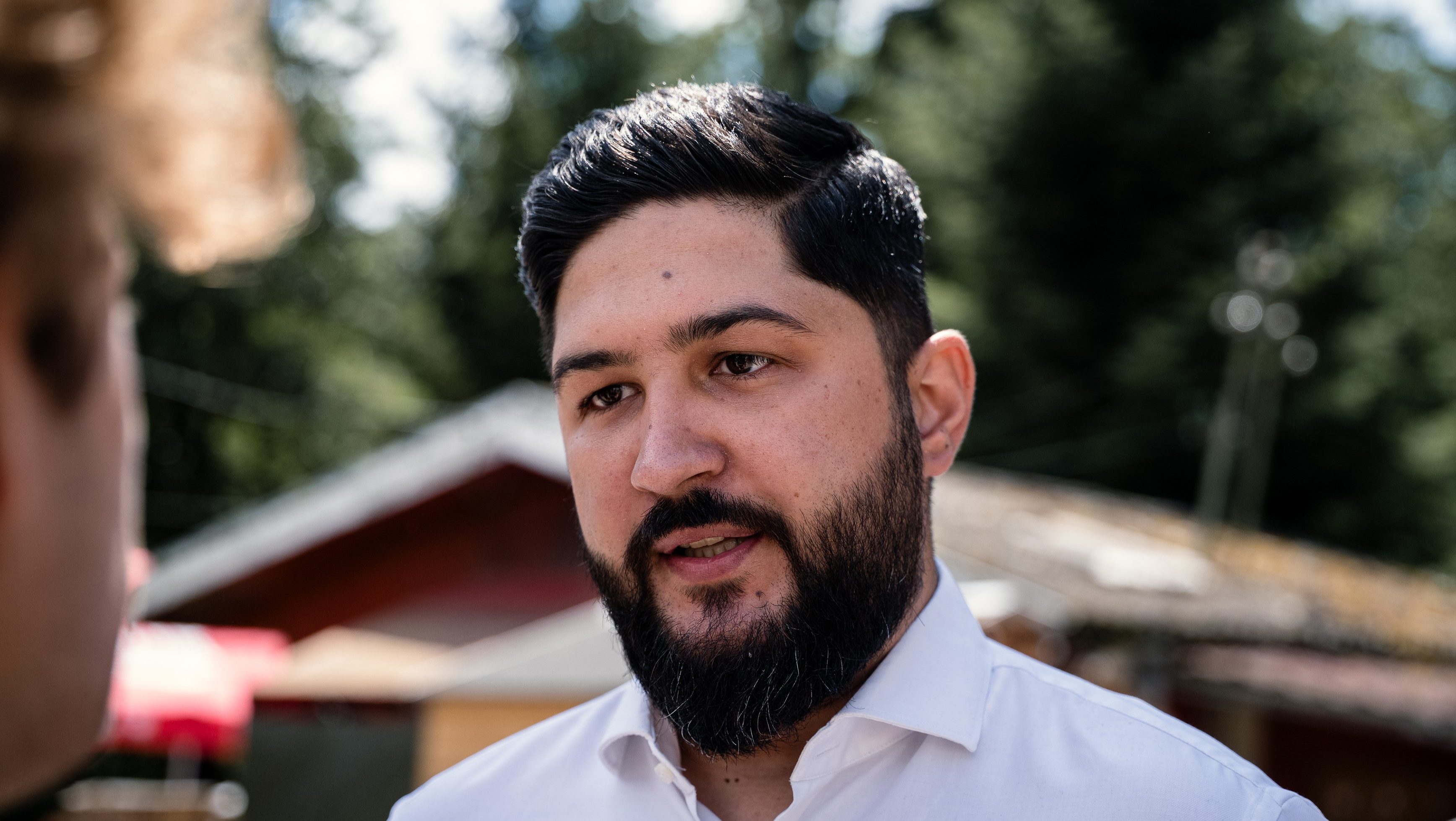 Man with dark hair and full beard wearing a white shirt, speaking outdoors to someone off-camera.