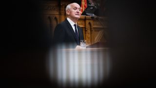 Older man in a dark suit speaks at a wooden lectern in a formal chamber, with a glass of water nearby and a microphone present.