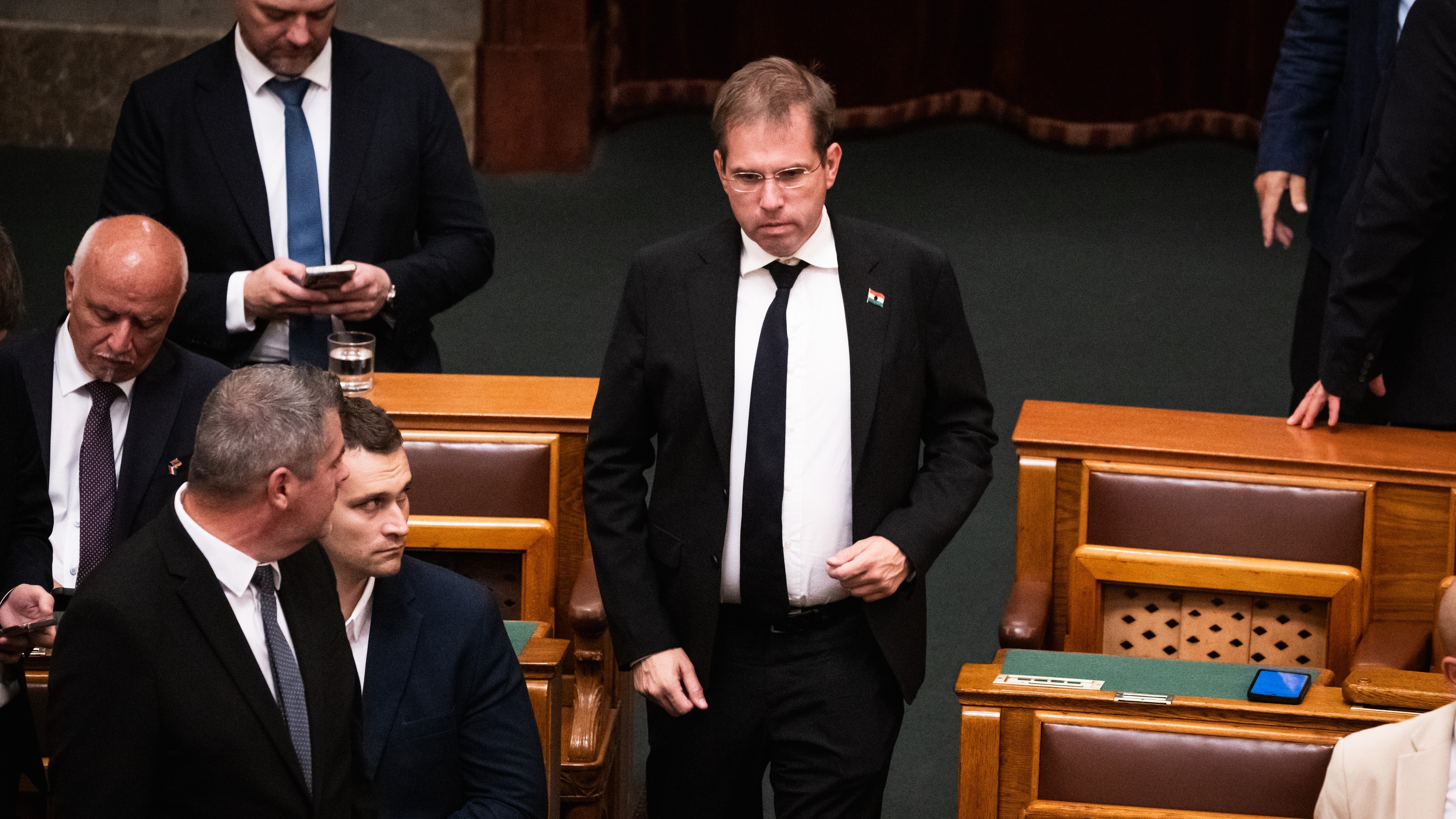 Man in a dark suit stands at a desk in a formal parliamentary chamber, surrounded by seated officials.