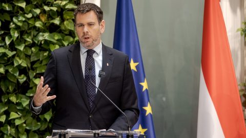 Man in a dark suit and tie speaks at a transparent lectern with microphones, EU and red-white flags in the background.