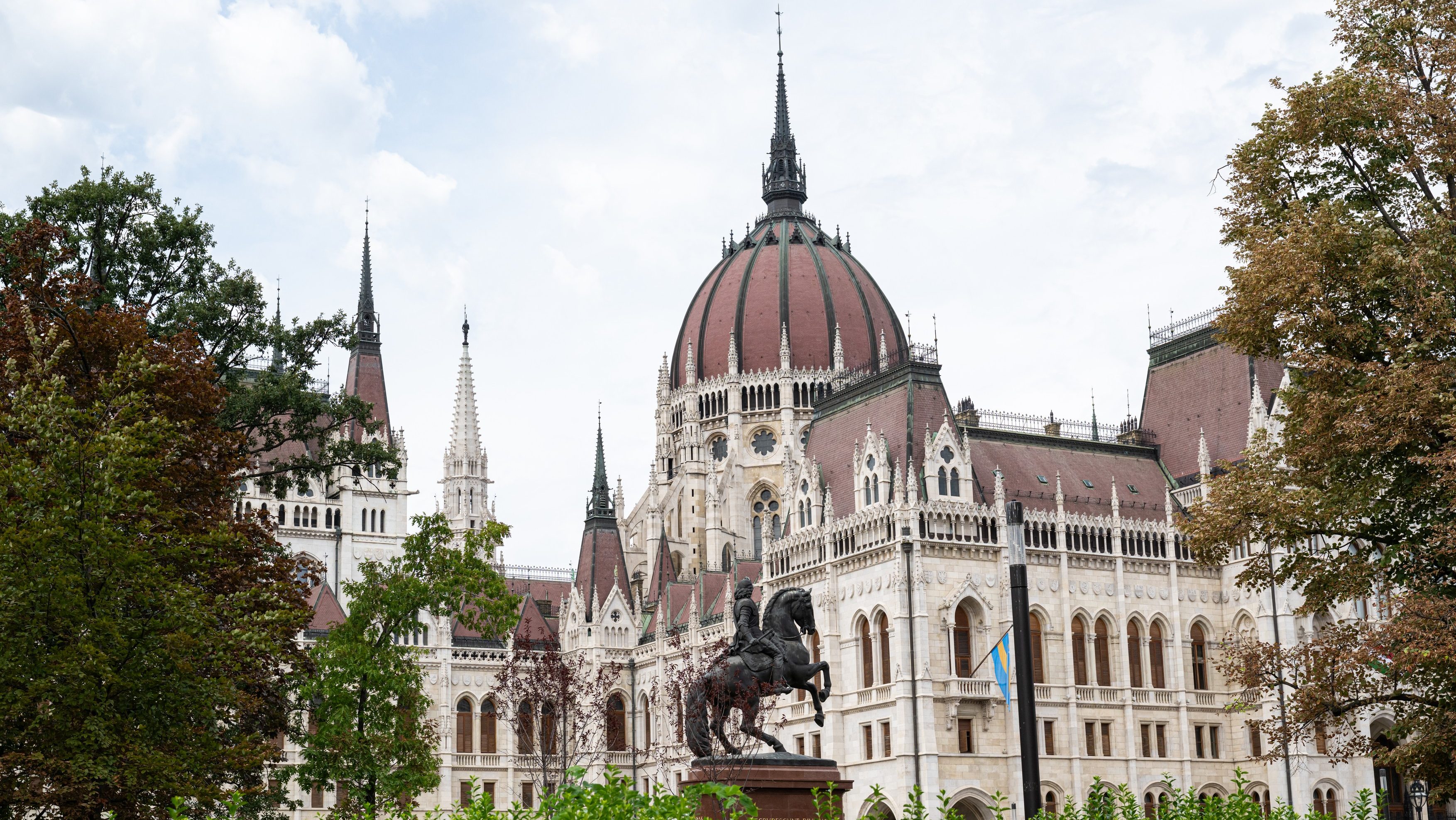 Budapest's Hungarian Parliament Building with a red dome and tall spires, framed by green trees in the foreground.