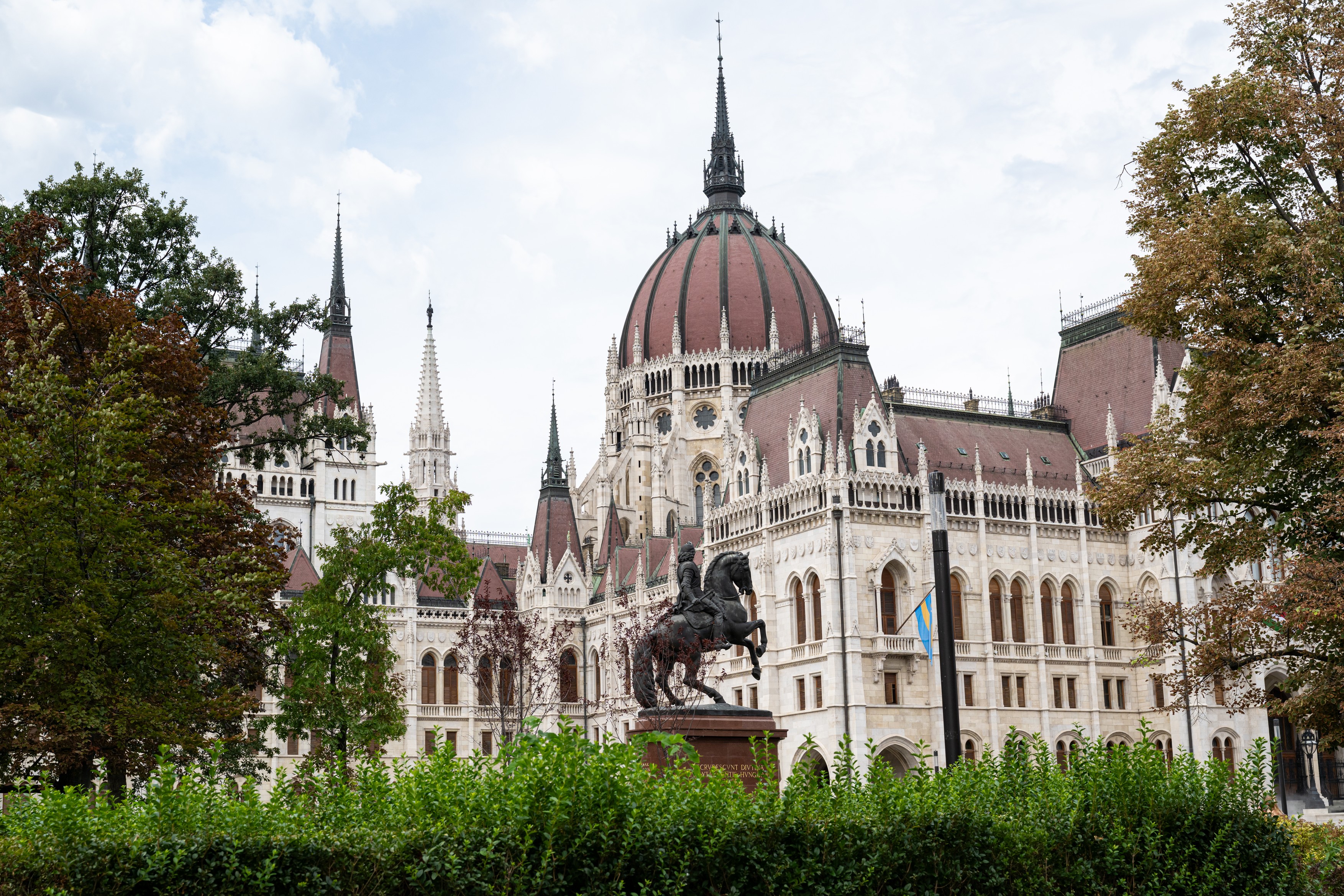 Budapest's Hungarian Parliament Building with a red dome and tall spires, framed by green trees in the foreground.