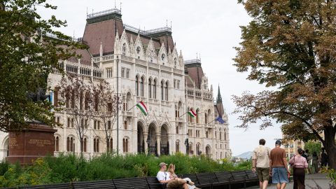 Ornate white Gothic-style building with red roofs, Hungarian flags, and people strolling in a park.