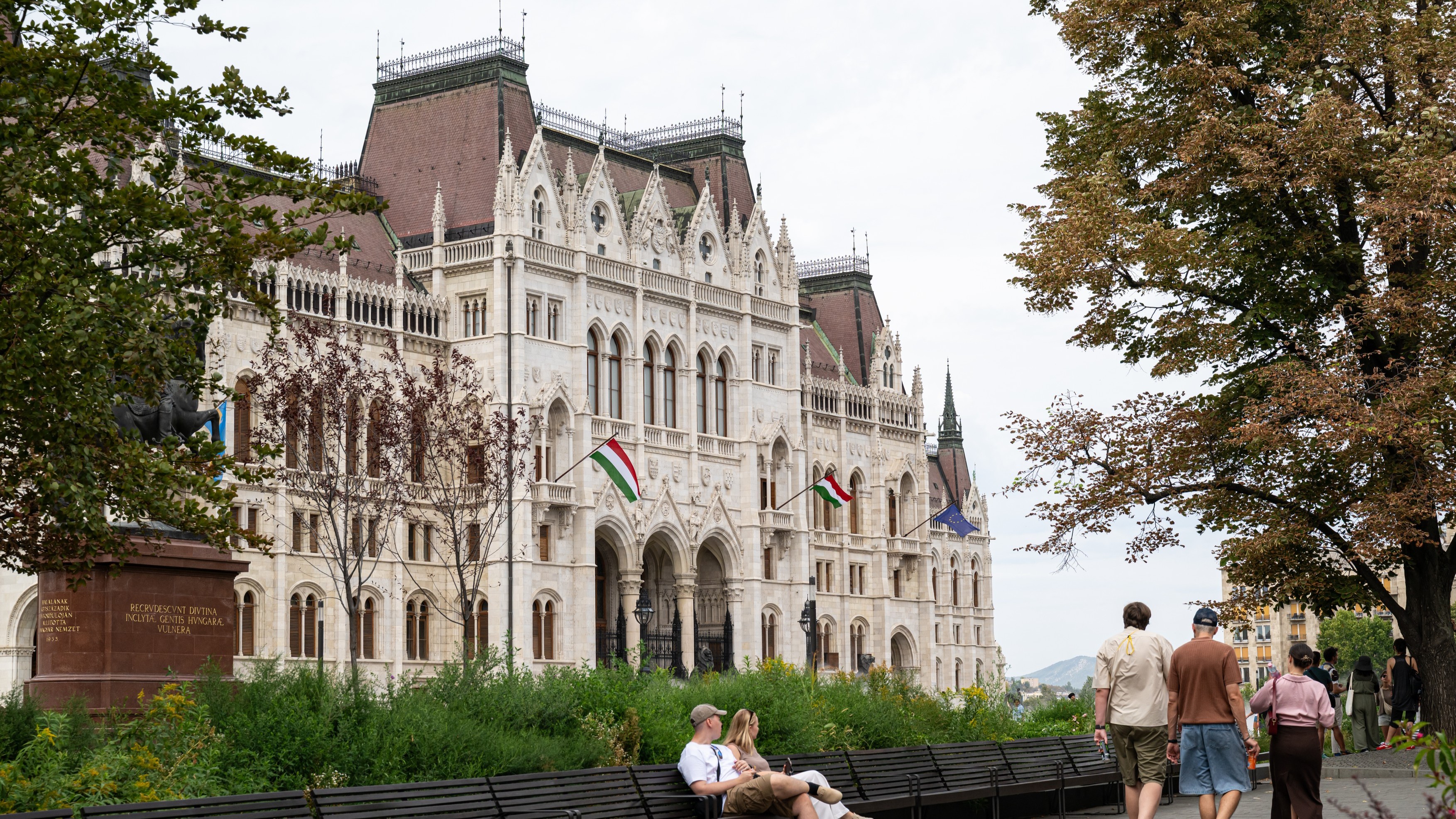 Ornate white Gothic-style building with red roofs, Hungarian flags, and people strolling in a park.