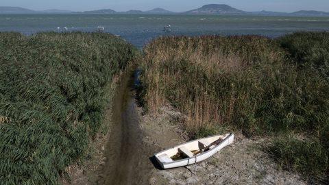 Narrow muddy water channel between tall reeds leading to a lake, with a small white boat on the shore and distant hills across the water.