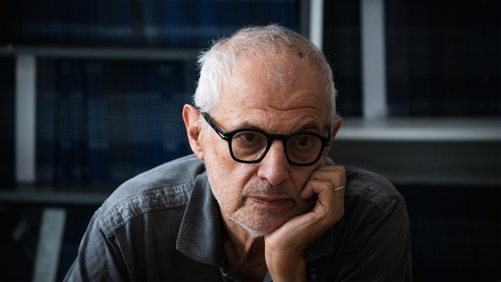 Older man with gray hair and glasses rests his chin on his hand in a dimly lit room, looking thoughtful.