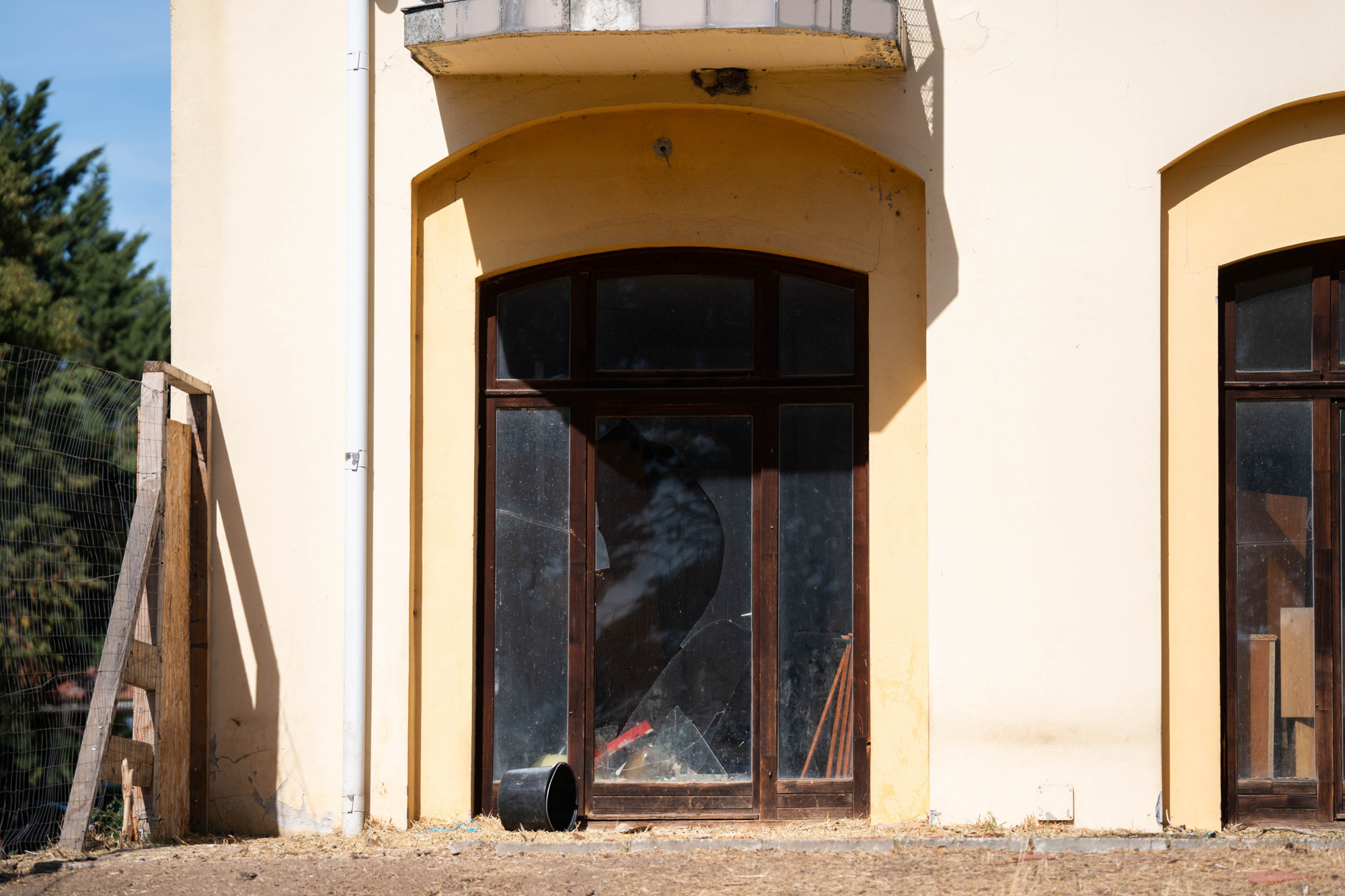 Beige building facade with a tall arched glass door, cracked panes, and a downspout along the left edge.