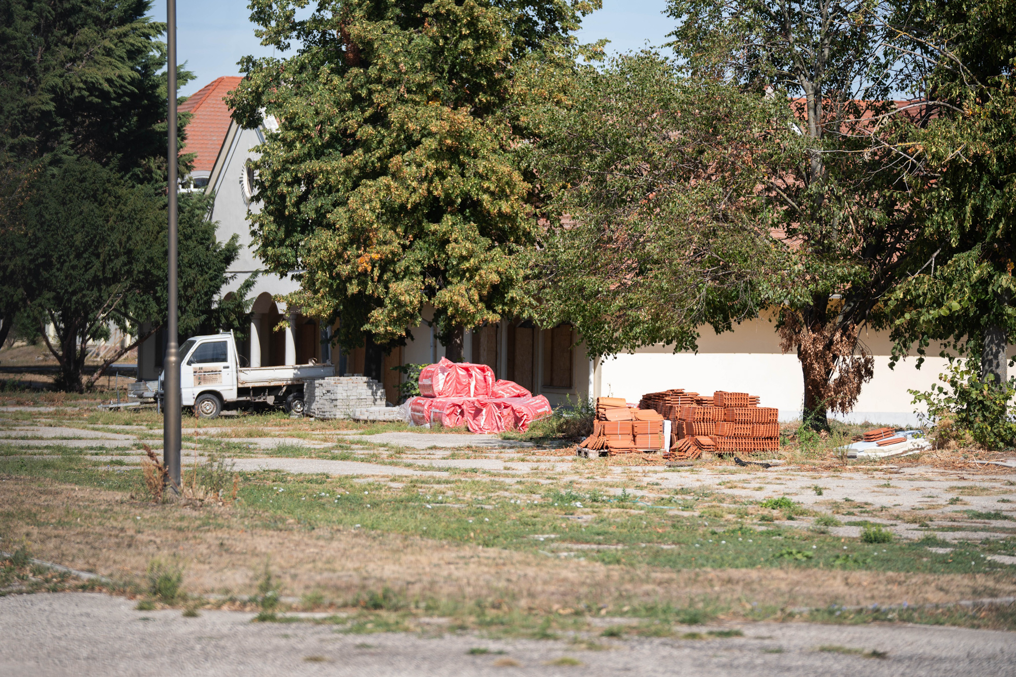 Construction site with stacked bricks, orange-wrapped bundles, and a small white truck beside a beige building under trees