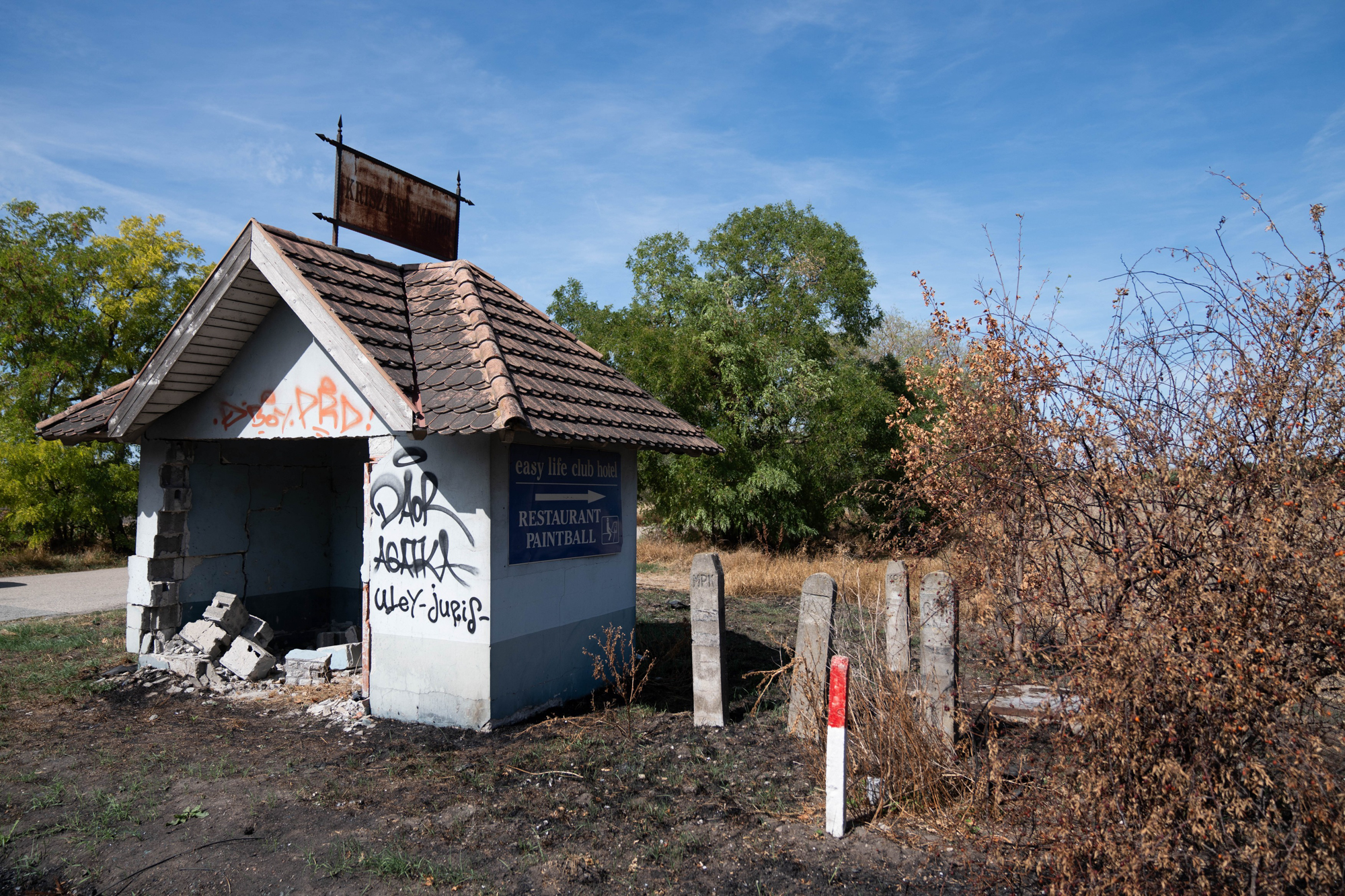 Abandoned small building with tiled roof, graffiti on walls, and a rusted sign against a clear blue sky.