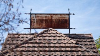 Rusty metal sign reading 'KRISZTINA-MAJOR' mounted on a wrought-iron frame above a tiled roof against a blue sky.