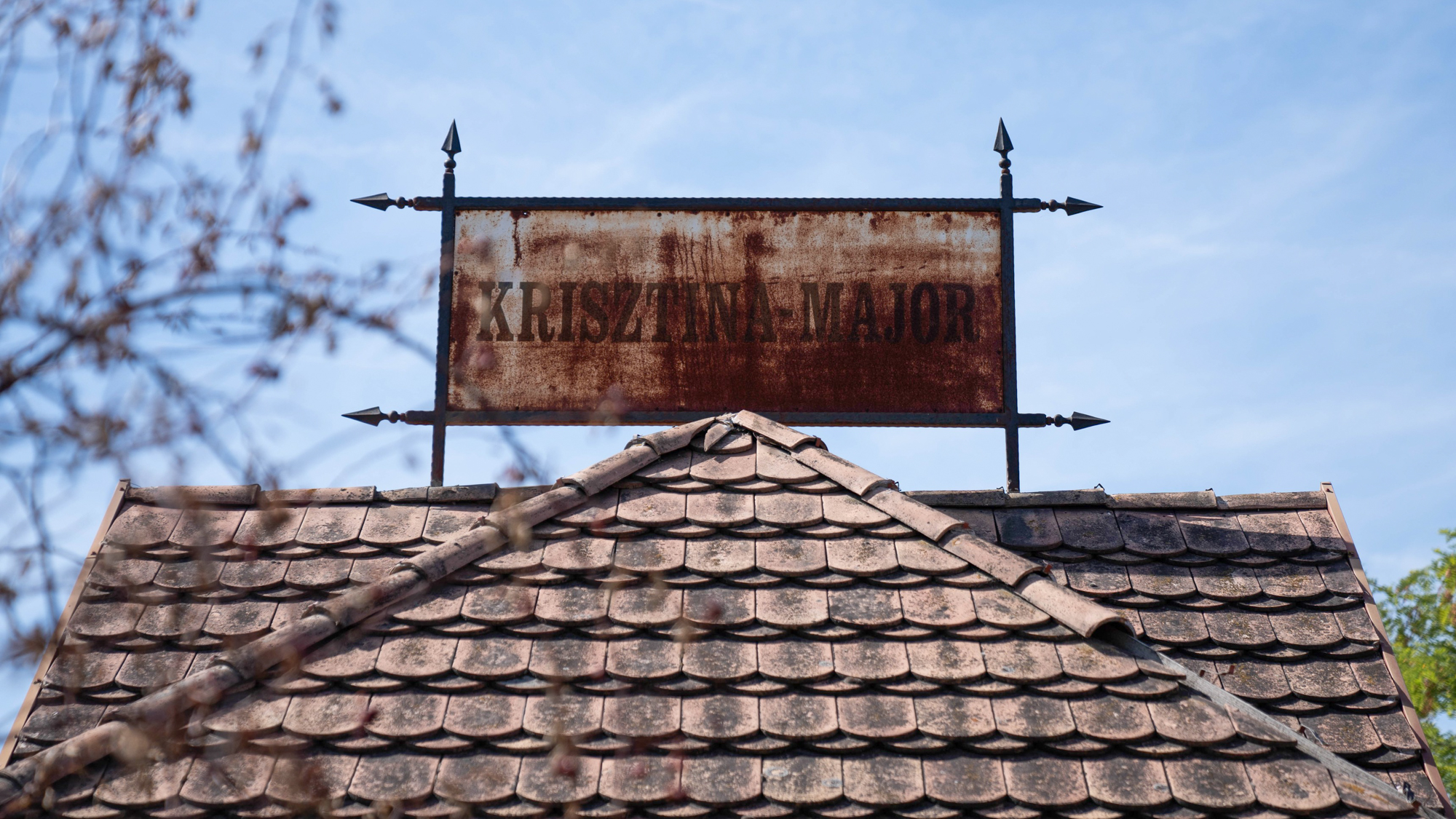 Rusty metal sign reading 'KRISZTINA-MAJOR' mounted on a wrought-iron frame above a tiled roof against a blue sky.