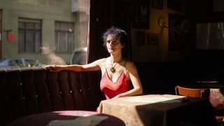 Woman in a red sleeveless top sits at a sunlit booth in a dim cafe, left arm resting on tufted upholstery.