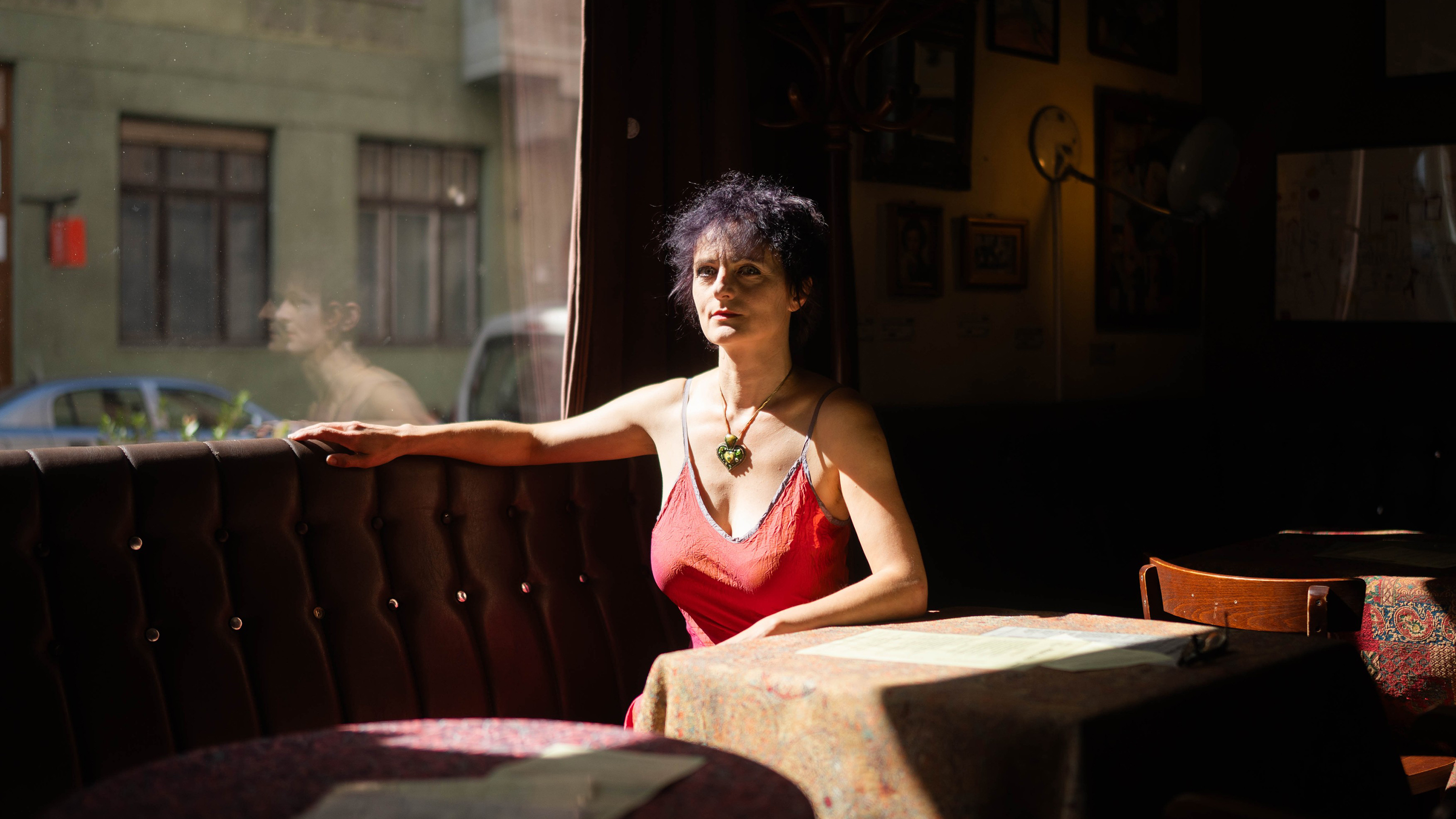 Woman in a red sleeveless top sits at a sunlit booth in a dim cafe, left arm resting on tufted upholstery.