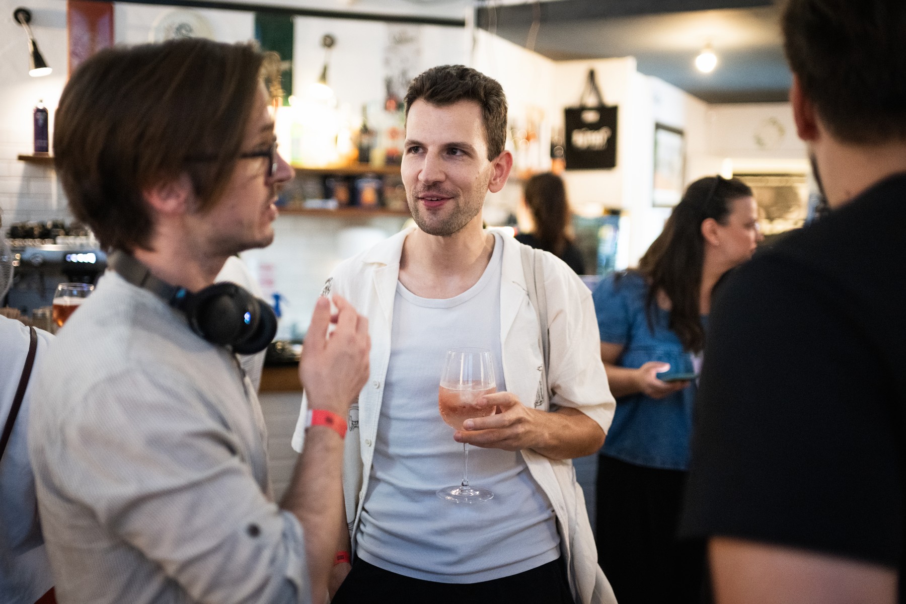 Man in a white shirt holding a wine glass chats with a friend wearing headphones at a bar social event, blurred background.