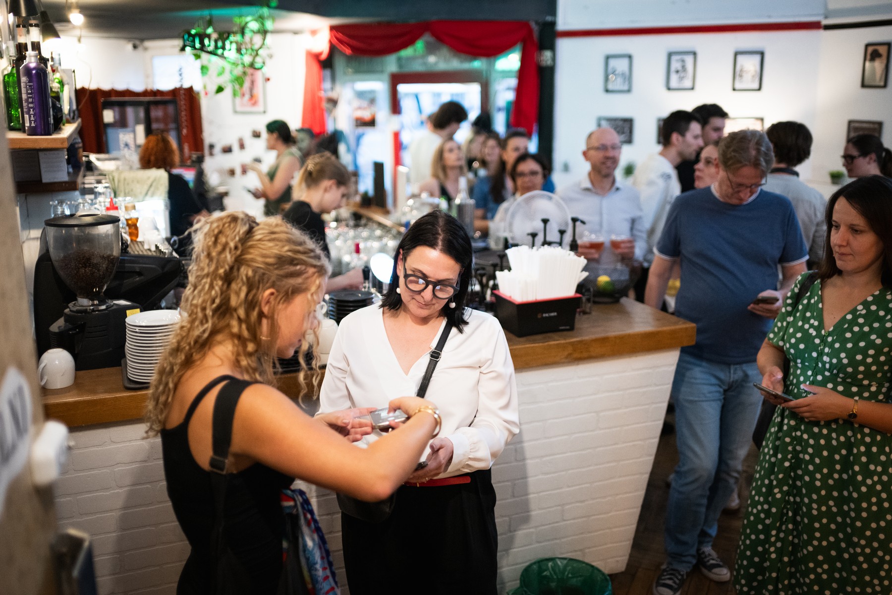 Two women at a bar counter exchanging a card or item while a crowded crowd mingles in the background.