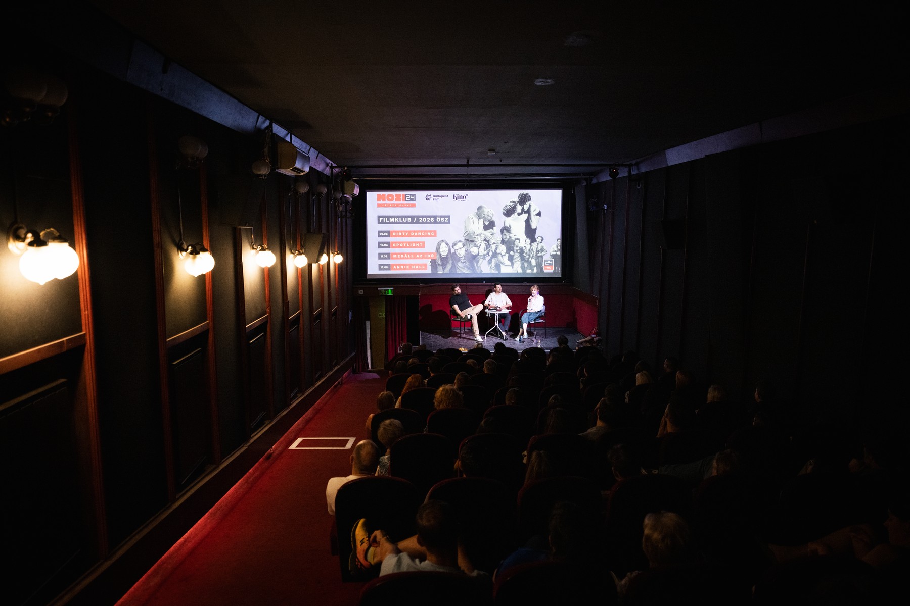 Dimly lit small theater with a panel of three people seated on stage in front of a large screen displaying a poster and audience watching from red-carpeted seats.