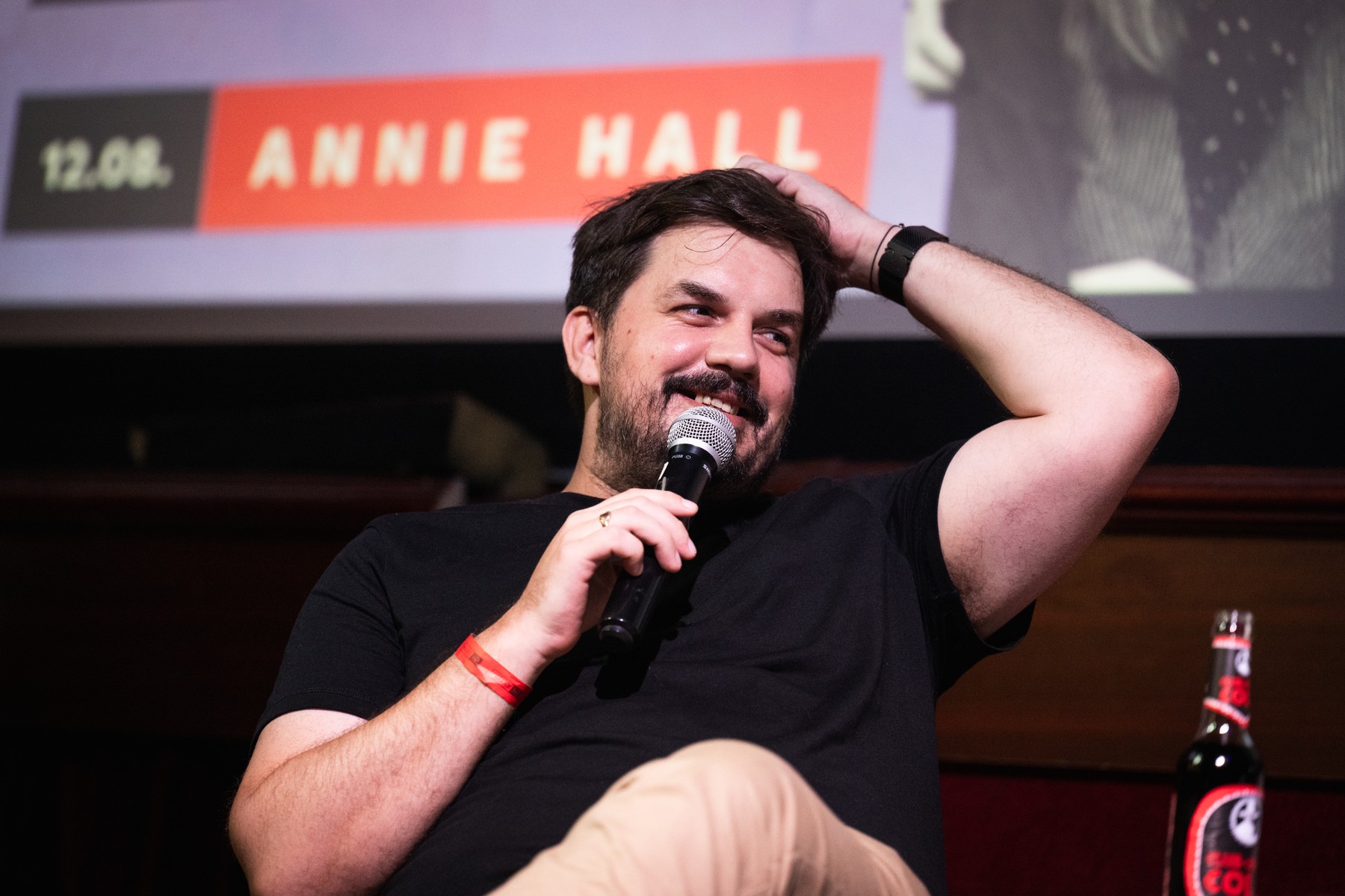 Man with dark hair and stubble, wearing a black T-shirt, smiling as he holds a microphone on stage.