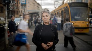 Portrait of a woman with short gray hair and colorful earrings standing on a busy city street, a yellow tram in the background.