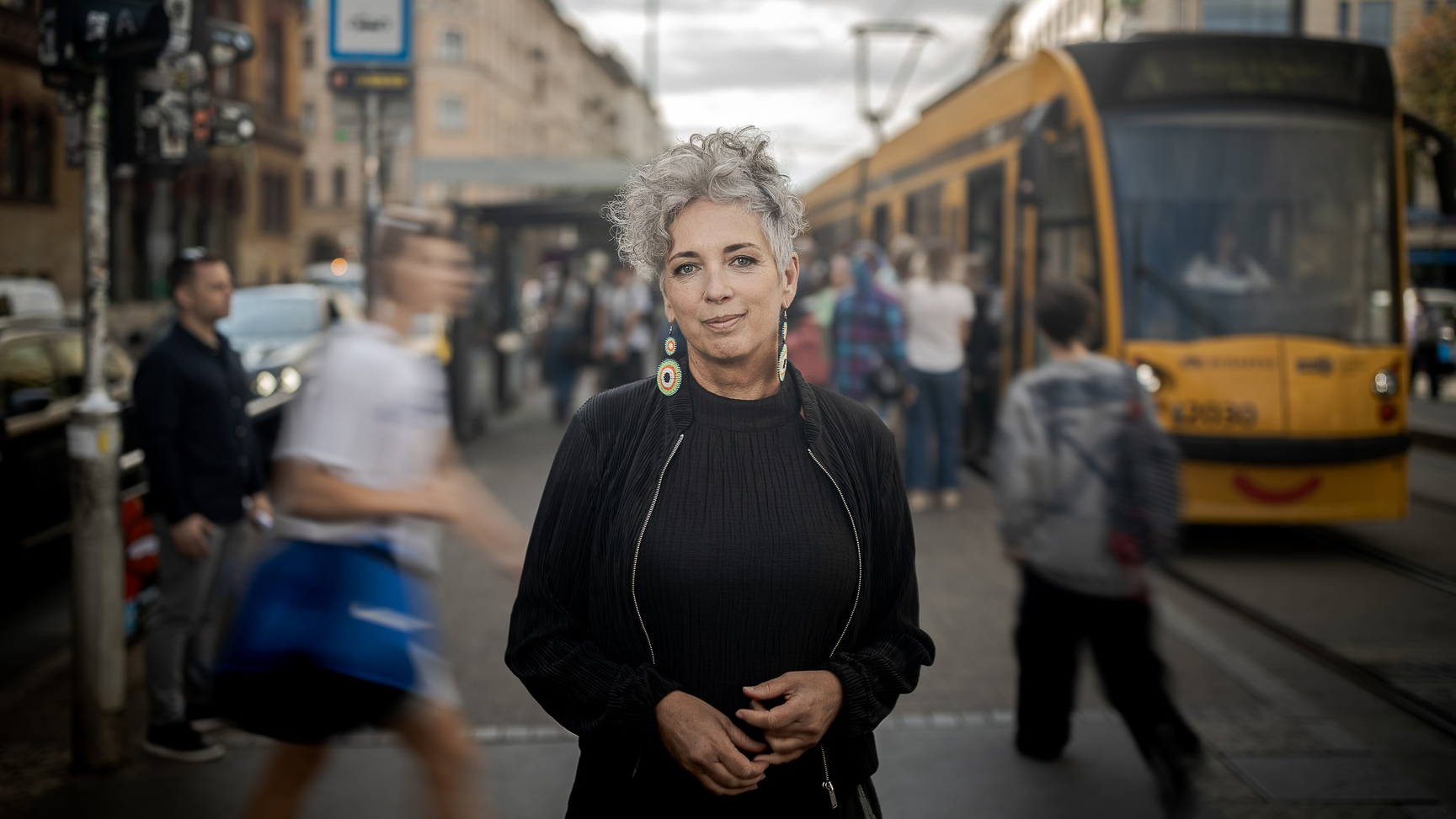 Portrait of a woman with short gray hair and colorful earrings standing on a busy city street, a yellow tram in the background.
