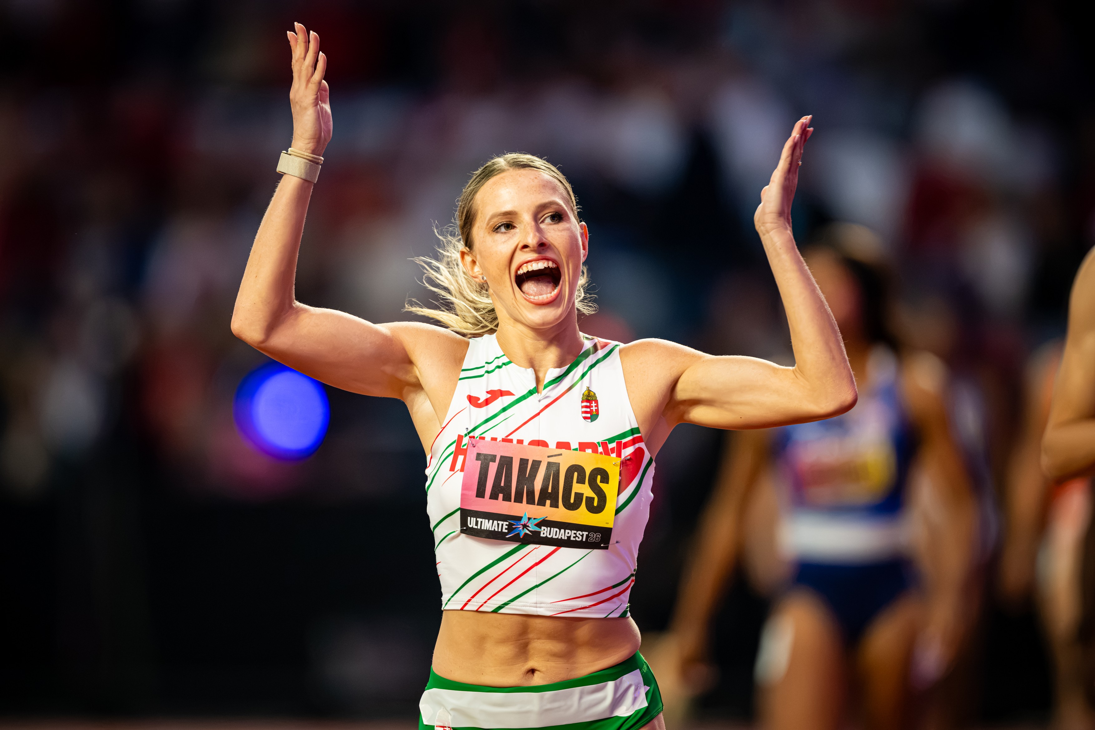 Athlete with arms raised in celebration, wearing a white jersey with Hungarian flag and 'TAKACS' bib badge at a race.