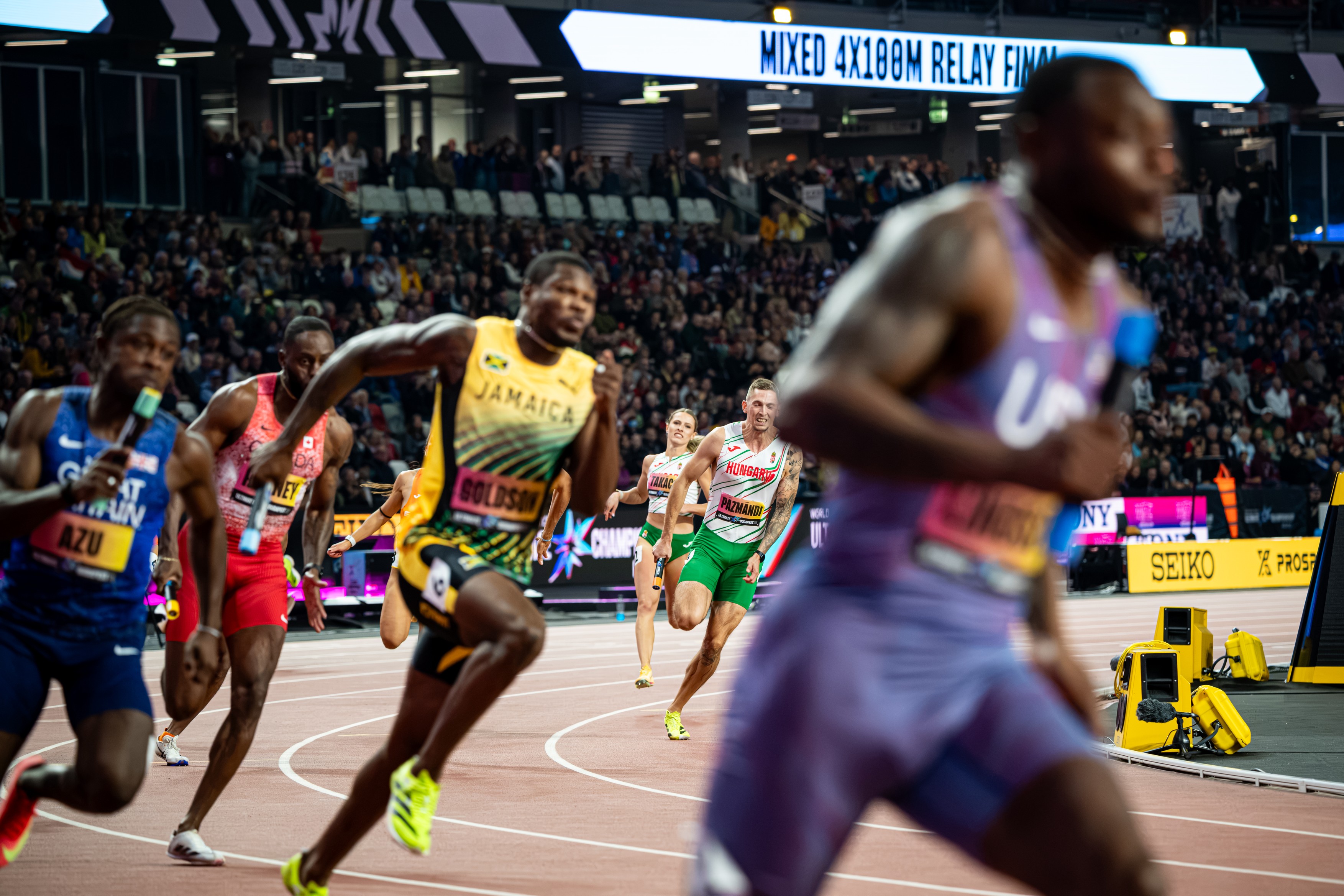 Professional sprinters race on a curved track with a Jamaican athlete in yellow leading, packed crowd in the stands behind them.