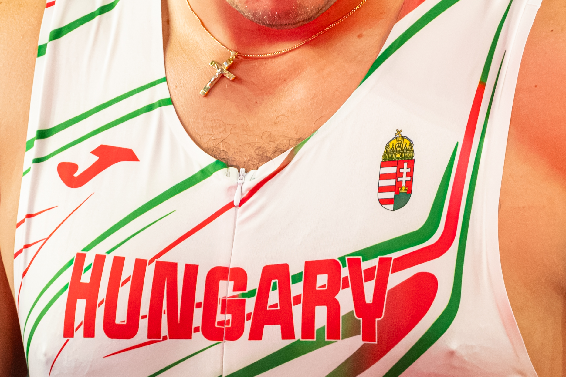 Close-up of a white sports jersey with green and red stripes, a Hungarian coat-of-arms emblem, and a gold cross necklace.