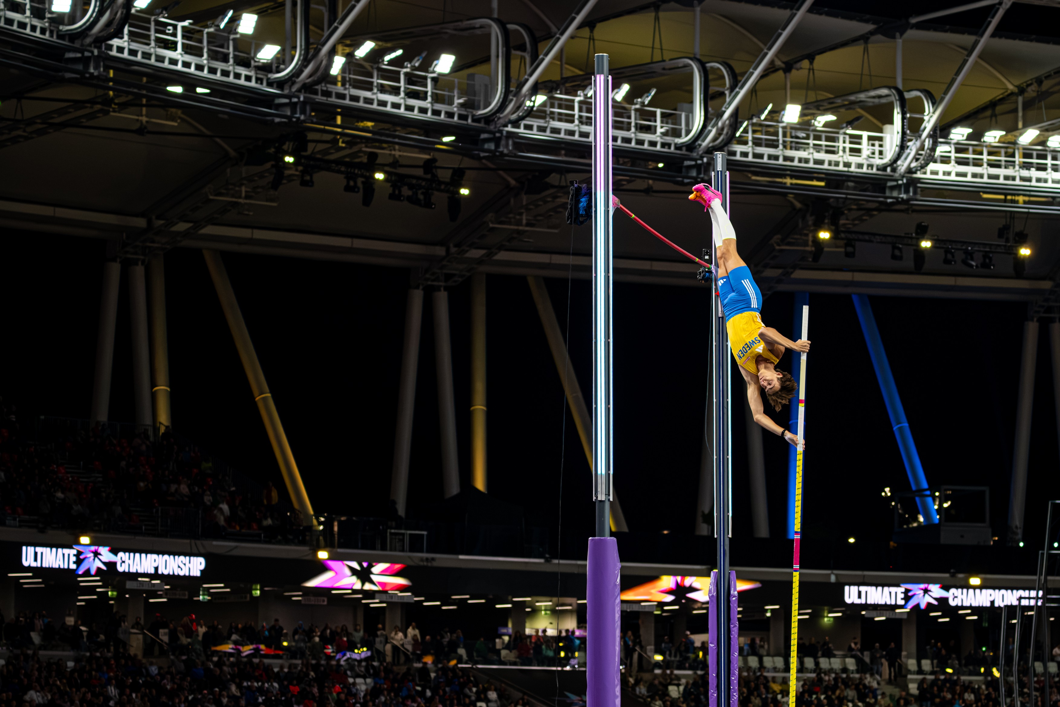 Pole vaulter in yellow shorts and blue top upside down on the pole during an indoor competition, with purple padding and a cheering crowd and 'Ultimate Championship' signage in the background