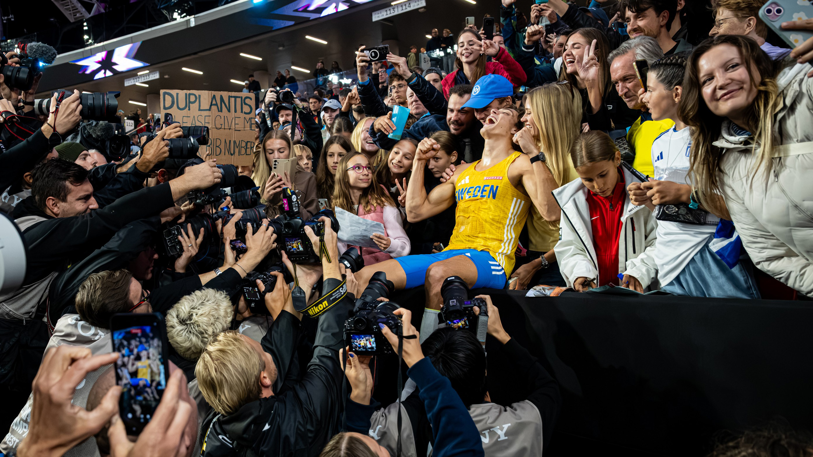 Swedish runner in a yellow Sweden kit celebrating while photographers crowd around him at a press event.