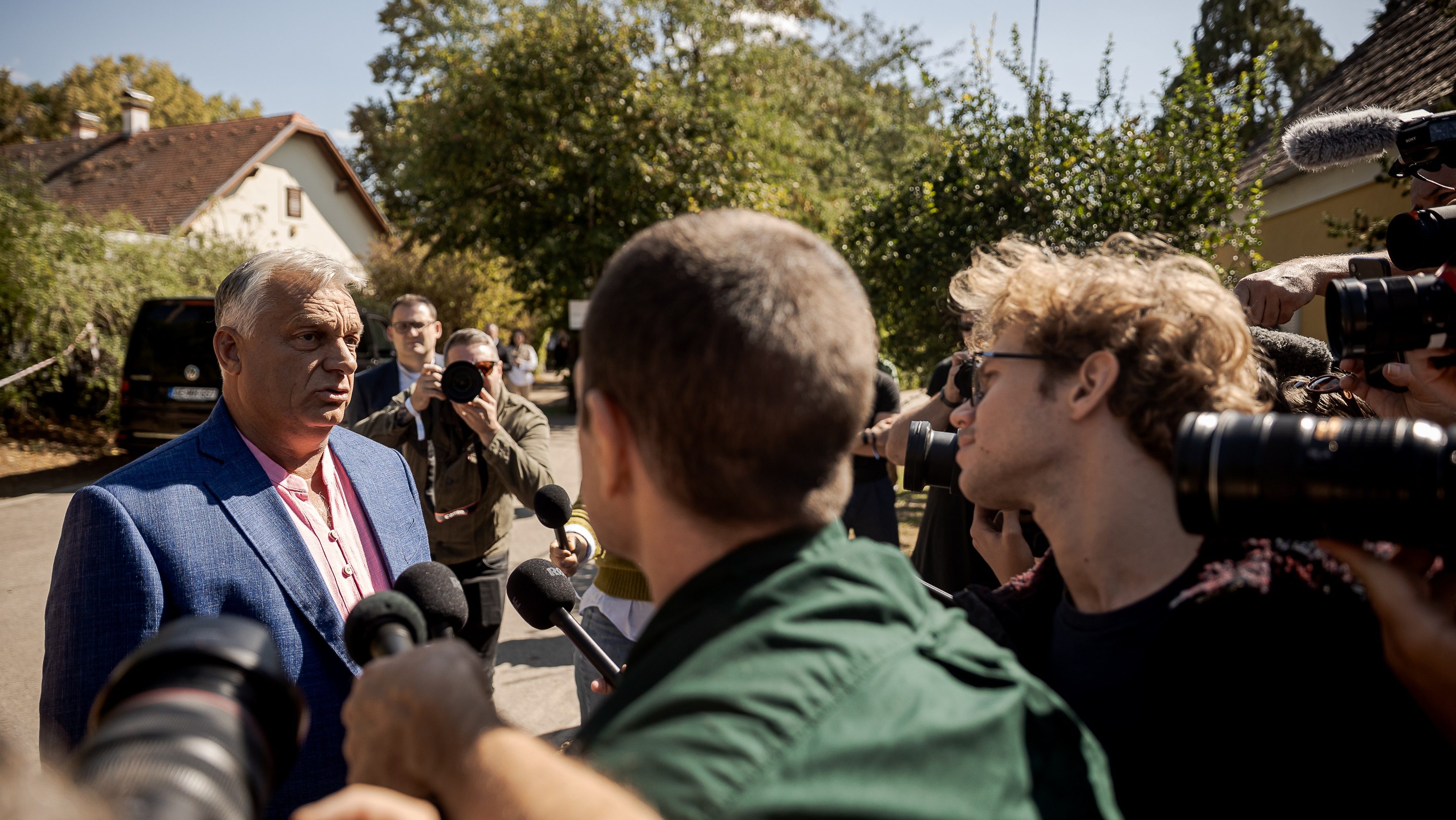 Man in a blue blazer being interviewed outdoors as several reporters point microphones and cameras toward him.
