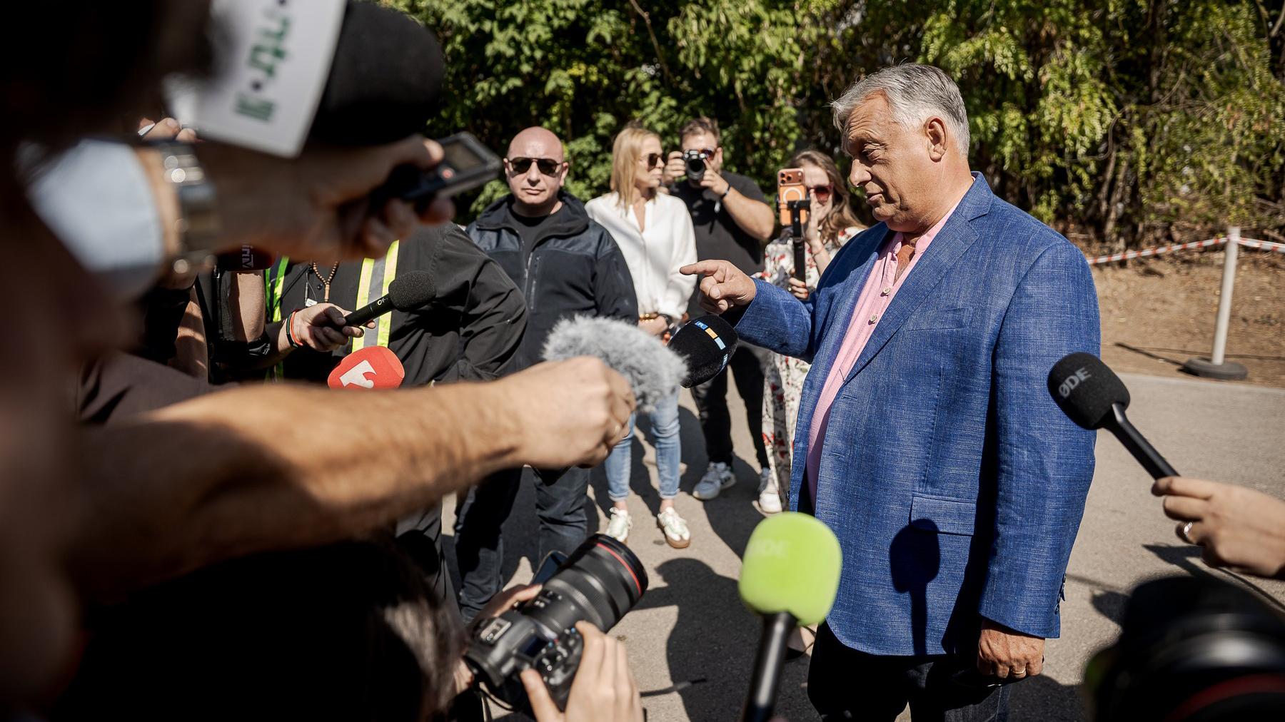 Older man in a blue blazer speaks to a crowd of reporters holding microphones outdoors.