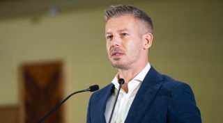 Male speaker in a suit delivering a presentation at a podium with dual microphones in a conference room.