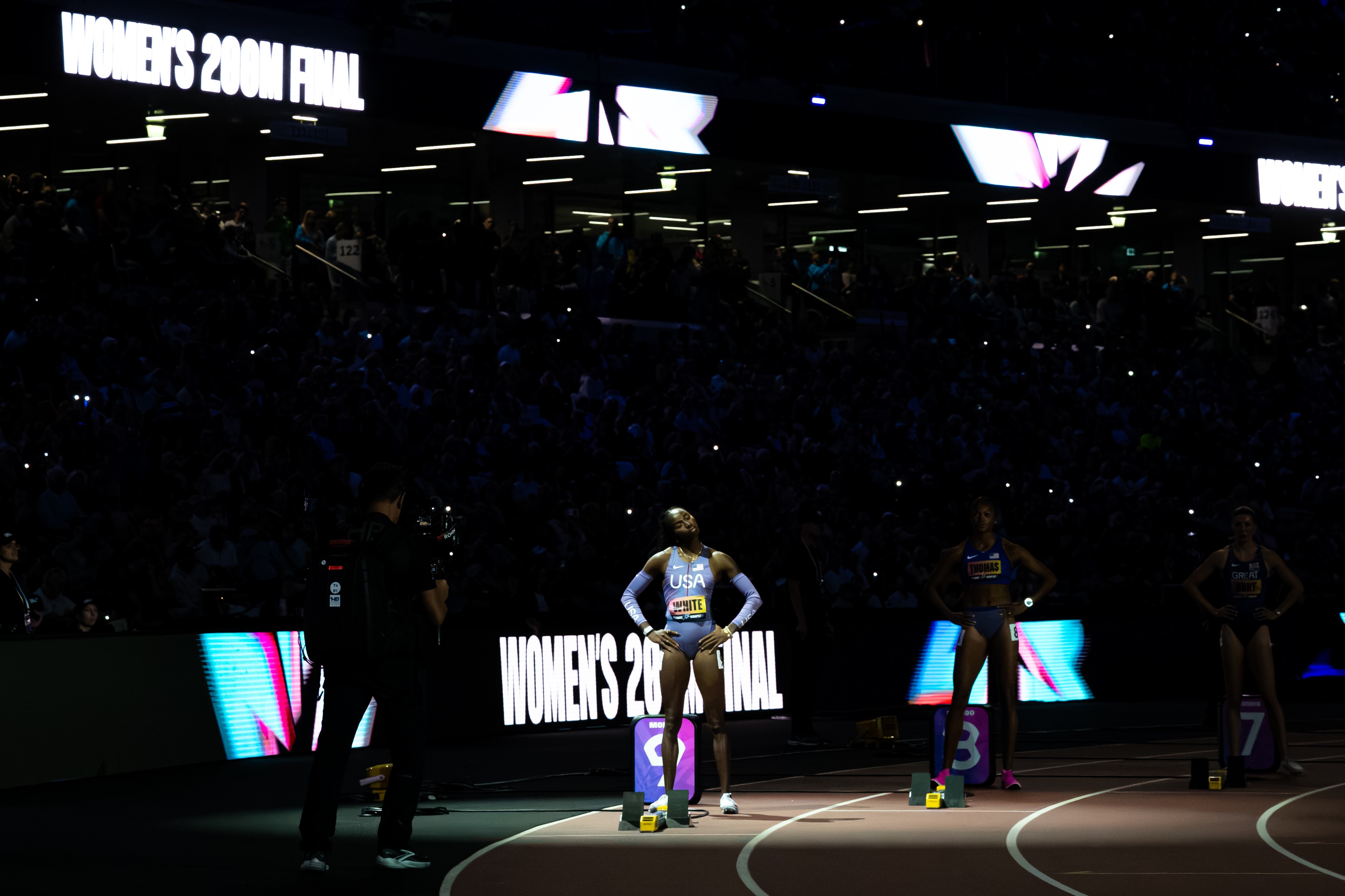 Athletes stand on starting blocks on a dim indoor track as the women's 200m final begins, with a large audience and bright signage reading 'WOMEN'S 200M FINAL'.
