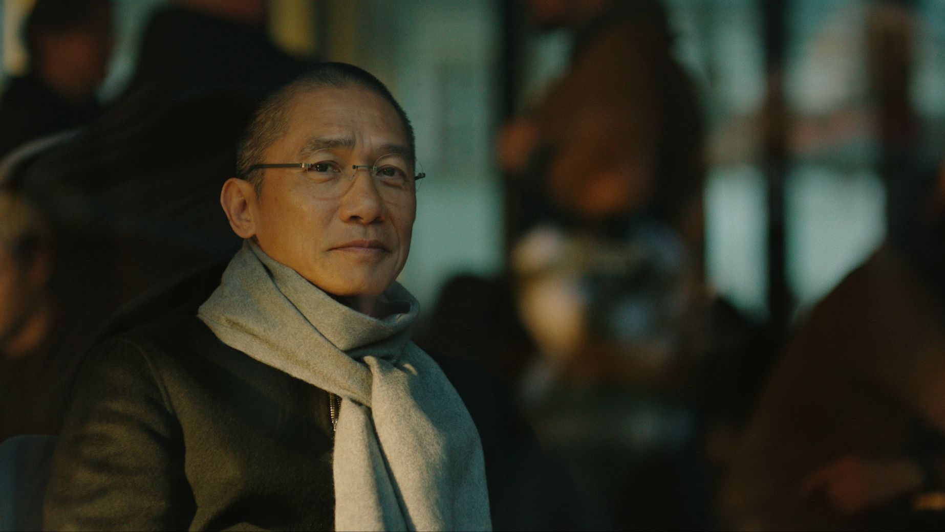 Portrait of a man wearing glasses and a light scarf, looking calmly toward the camera in a dimly lit setting.