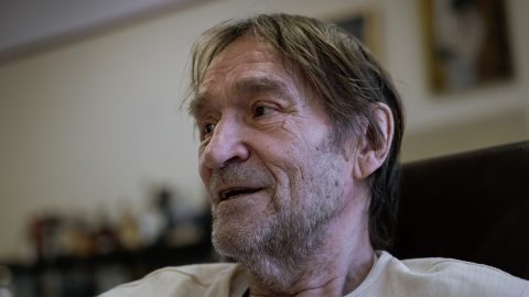 Close-up of an older man with a gray beard and light hair, looking to the left indoors.