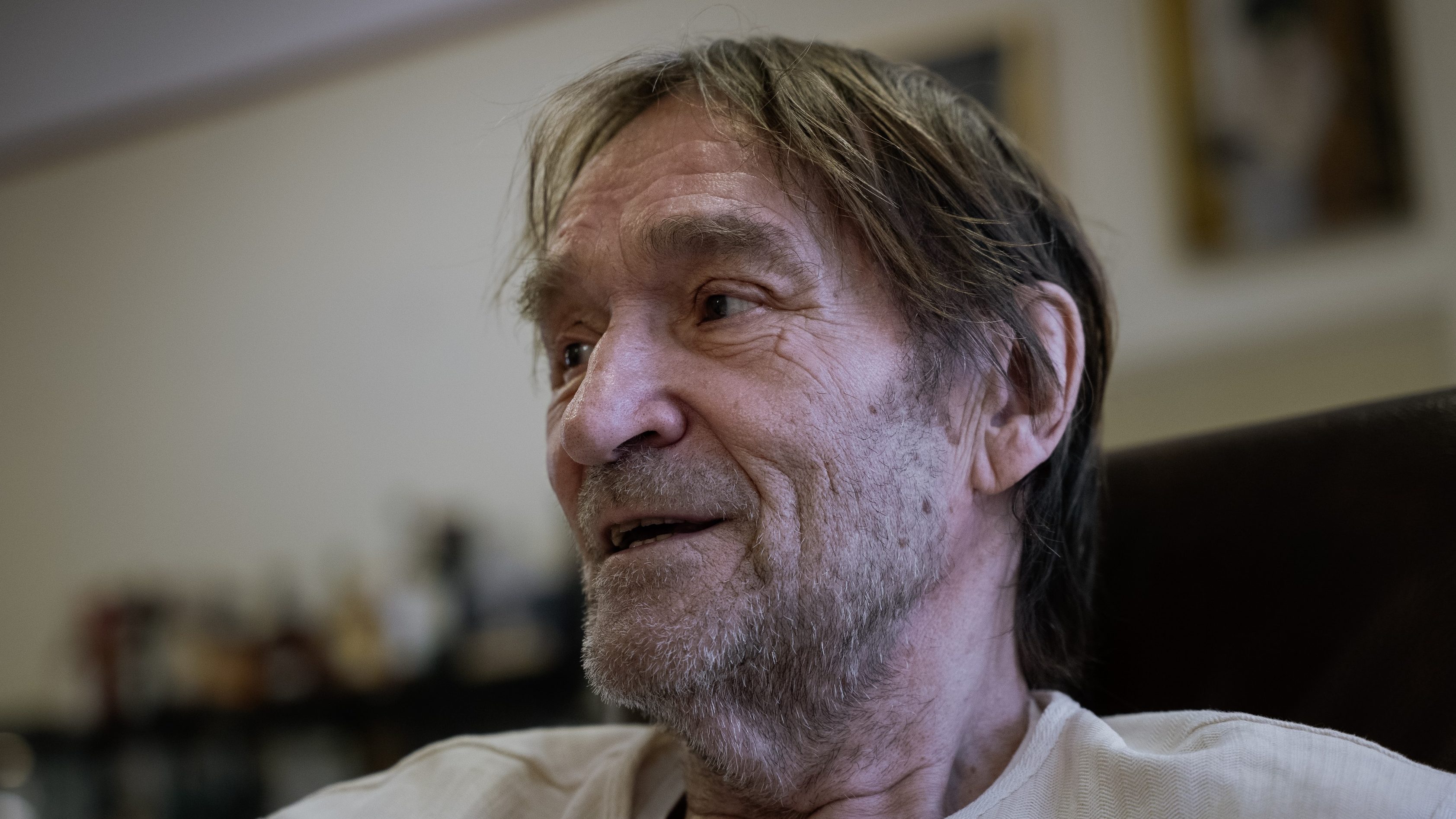 Close-up of an older man with a gray beard and light hair, looking to the left indoors.