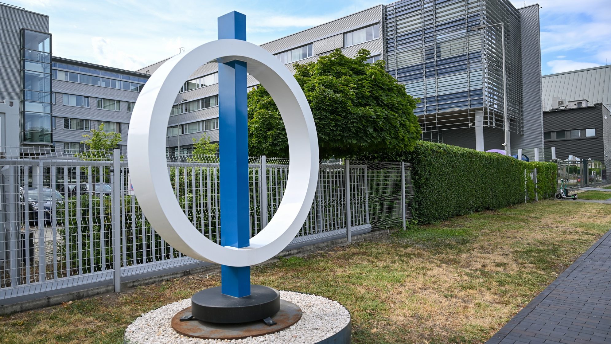 Urban office complex with a large white circular sculpture on a blue supporting post in a fenced courtyard.
