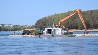 Orange excavator on a floating barge extends its arm to a red clamshell bucket dredging near a rocky shoreline; two workers stand on the water's edge with trucks nearby and a tree-covered bank in the background.