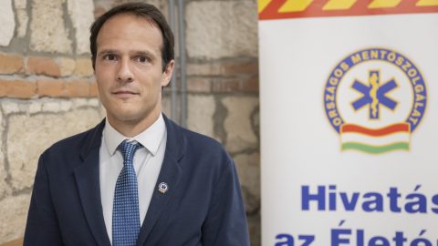 Man in a navy suit and blue tie posing beside a banner with a medical symbol, brick wall behind him (health event).