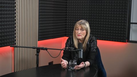 Woman in a black outfit sits at a dark table in a recording studio, speaking into a microphone with acoustic foam panels behind her and a glass of water on the desk
