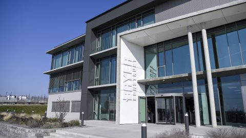 Modern office building exterior with a glass façade, gray panels, and prominent white entrance panel under blue sky.