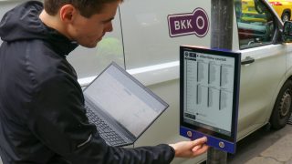Person in a dark jacket uses a laptop while standing next to a bus-timetable display at a street stop, near a white van with a BKK sign.