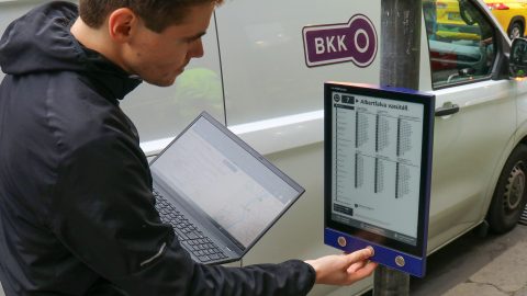 Person in a dark jacket uses a laptop while standing next to a bus-timetable display at a street stop, near a white van with a BKK sign.