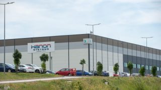 Exterior view of a large industrial warehouse with a Hannon Systems sign, a row of parked cars, and trees in the landscaping under a blue sky.